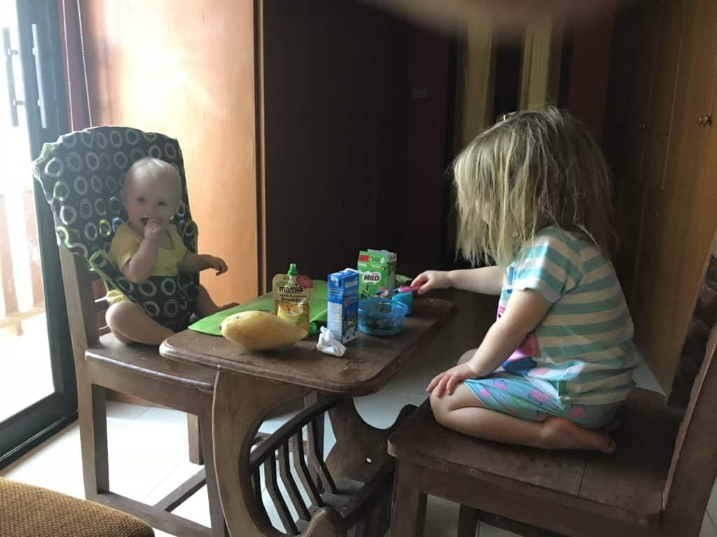 Baby in totseat chair harness and toddler eat breakfast in bedroom in Thailand