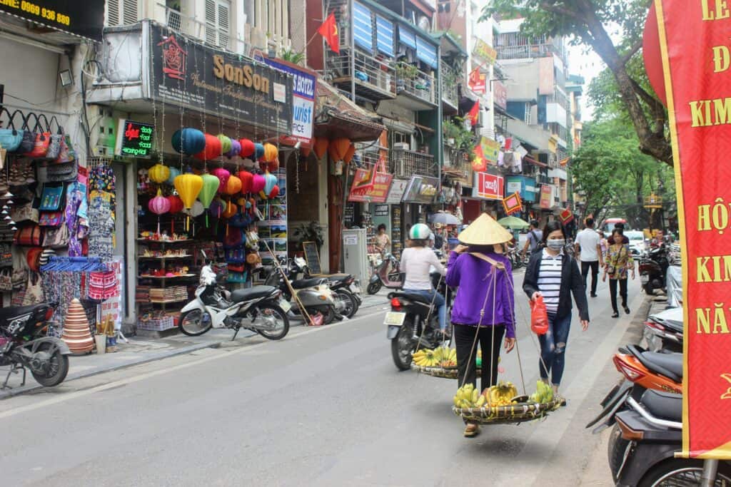 Seller in traditional veitnames hat walks a busy street in Hanoi with kids